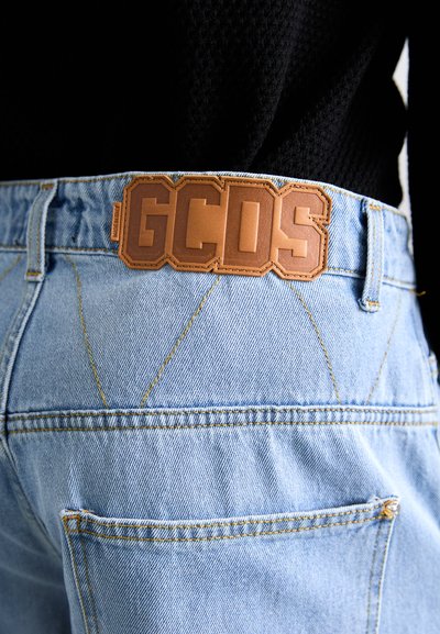 Vaqueros de denim azul claro con costuras visibles, presentan una etiqueta de cuero marrón con el logo en relieve "GCDS" en la cinturilla.