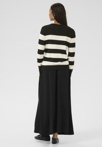 Pull rayé noir et blanc à manches longues, jupe noire et chaussures plates noires, vu de dos sur un fond clair.