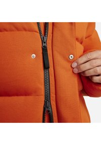 Orangene Puffjacke mit einem schwarzen Reißverschluss und Druckknöpfen. Das Material wirkt strukturiert und hat ein gepolstertes Design zur Isolierung.