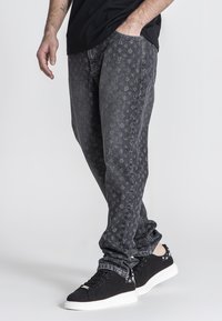 Jeans de mezclilla negros con un patrón de logotipos en toda la prenda, diseño ajustado y un acabado ligeramente desgastado, combinados con zapatillas negras.