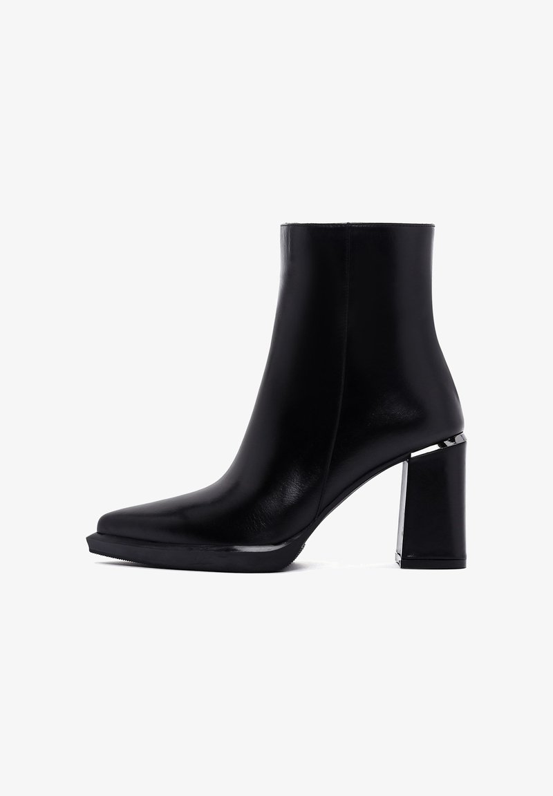 Botte de cheville noire en cuir lisse avec un bout pointu. Présente un talon carré épais et des accents argentés le long de la base.