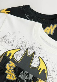 Svart och vit sweatshirt med en gul Batman-logotyp och spruttryck. Sträckbar krage och synlig texturerad tyg.