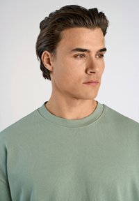 Jasnozielona, prążkowana bluza z okrągłym dekoltem i fakturowanym materiałem, noszona przez modelkę o krótkich, ciemnych włosach, lekko zwróconą w bok.