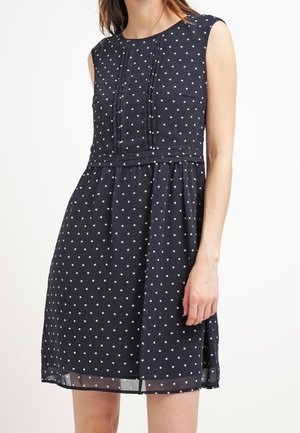 Granatowa sukienka bez rękawów w białe grochy, z plisowanym przodem, dopasowaną talią i spódnicą do kolan, noszona przez stojącą osobę.