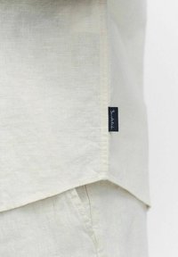 Lichtbeige linnen shirt met een afgeronde zoom en smalle zijsplitten, voorzien van een klein donkerblauw merklabel dat in de zijnaad is genaaid.