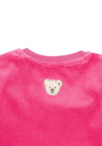 Prenda de velour rosa con un escote redondo, que presenta un aplique de cara de oso en blanco y negro justo encima del escote en la parte posterior.
