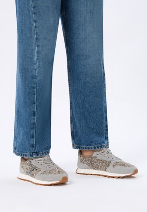 Person wearing blue denim jeans and gray textured sneakers with white soles and brown tread standing on a plain white background.