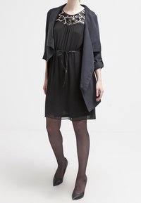 Robe noire avec un col en dentelle, taille cintrée avec un lien, tissu superposé. Portée avec une veste ouverte noire assortie et des chaussures à talons hauts noires.