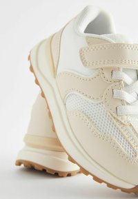 Beżowe buty sportowe z siateczkowym i syntetycznym wierzchem, teksturowaną podeszwą oraz regulowanym paskiem. Posiadają kontrastowe szwy i wyściełany kołnierz.