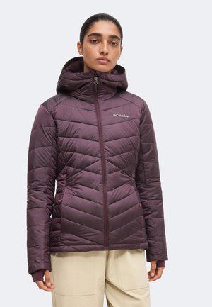 Kvinde iført en mørklilla quiltet Columbia jakke med hætte, stående med neutralt ansigtsudtryk og beige bukser mod en ensfarvet baggrund.