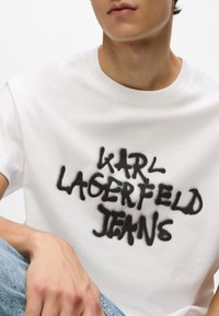Tânăr îmbrăcat cu un tricou alb pe care scrie "KARL LAGERFELD JEANS" în stil graffiti negru, asortat cu blugi albaștri.