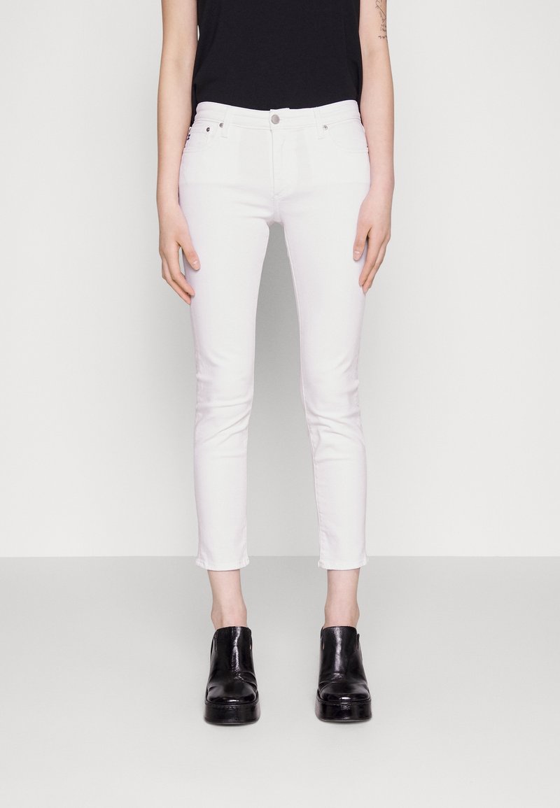Jeans blancos ajustados con una cintura de altura media, que cuentan con cinco bolsillos, costuras sutiles y un acabado limpio. Combinados con botas de tacón negras.