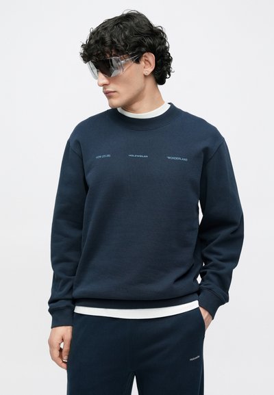 Granatowa bluza z okrągłym dekoltem, z białymi napisami. Do kompletu pasujące spodnie dresowe i przezroczyste okulary przeciwsłoneczne. Miękka struktura tkaniny.