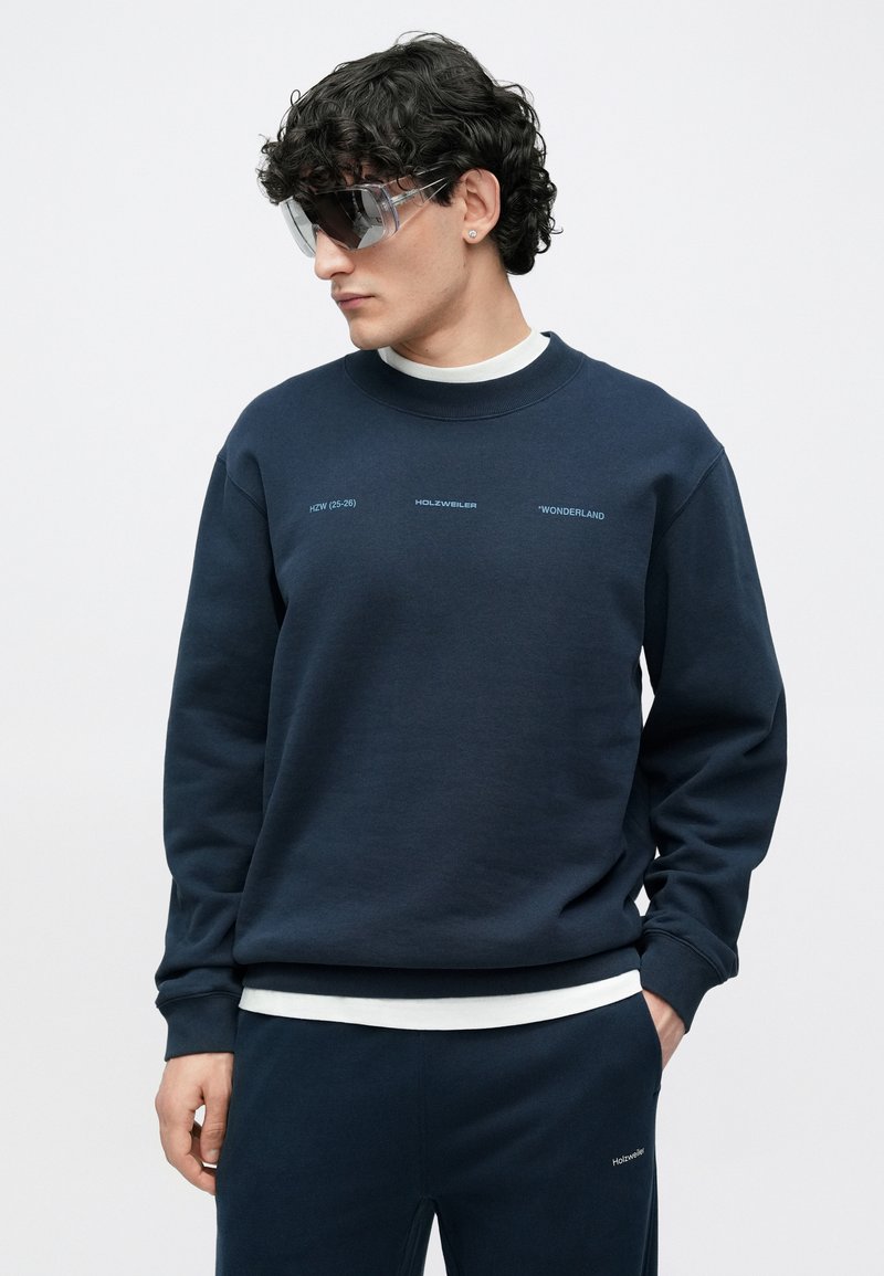 Sweat-shirt bleu marine à col rond, avec des détails en texte blanc. Associé à un pantalon de jogging assorti et des lunettes de soleil transparentes. Texture de tissu douce.