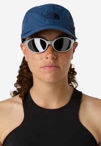 Blå keps med "The North Face"-logotyp, tillsammans med grå ovala solglasögon med reflekterande linser. Svart ärmlös topp. Lockigt hår synligt.