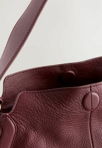 Sac à main en cuir texturé bordeaux foncé avec une large bandoulière et une fermeture à bouton pression rond, présenté ouvert sur un fond uni.