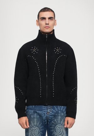 Black zip-up sweater made of soft knit fabric, featuring decorative studs arranged in curved patterns on the front and sleeves.