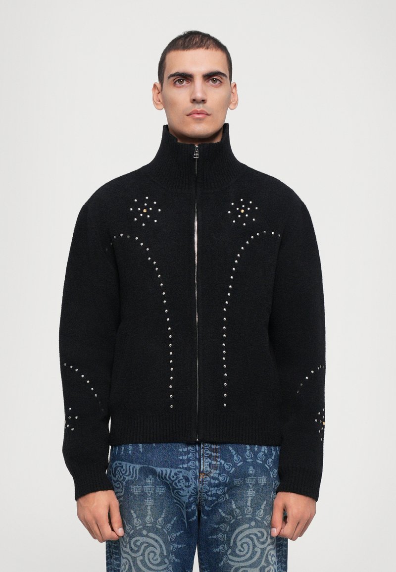 Črna zip-up jopica iz mehkega pletenega materiala, ki ima dekorativne žebljičke razporejene v ukrivljenih vzorcih na sprednji strani in rokavih.