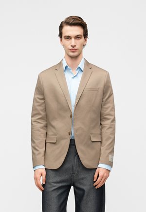 Mann trägt beige Blazer über hellblauem Hemd und dunkelgrauer Hose, steht vor weißem Hintergrund.
