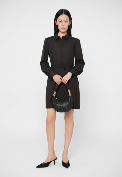 Sieviete, tērpusies melnā pogājamā kleitā ar garām piedurknēm, tur melnu rokassomu ar zelta akcentiem un vilk melnus smailos papēžus.