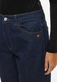 Mørkeblå denimjeans med gylden søm, med en hjerteformet detalj på lommen. Materialet har en glatt tekstur.