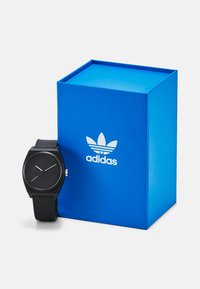 Montre analogique noire avec un bracelet en silicone texturé et un cadran noir minimaliste ; présentée à côté d'une boîte bleue avec le logo Adidas blanc.
