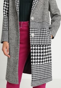 Schwarz-weißer Mantel mit Hahnentrittmuster und Fischgrätmuster, doppelt knopfbarer Schnitt mit silbernen Knöpfen, getragen über pinkfarbenen Hosen.