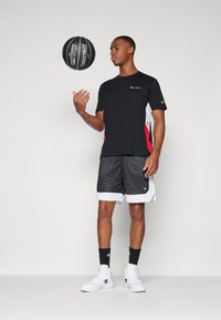 Camiseta deportiva negra con detalles en rojo y blanco, combinada con pantalones cortos de baloncesto de malla grises y zapatillas blancas, sosteniendo un balón de baloncesto negro.