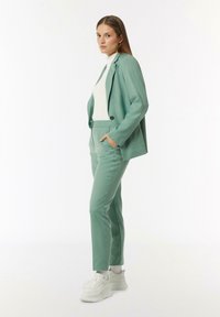 Hellgrüner Anzug mit einem maßgeschneiderten Blazer und passenden schmal geschnittenen Hosen, kombiniert mit einem weißen Rollkragenpullover und weißen Sneakern.
