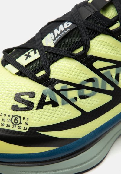 Κίτρινο και μαύρο sneaker MM6 Margiela με μαύρα κορδόνια, επάνω μέρος από δίχτυ, έντονη επιγραφή "SACAI" και τυπωμένους αριθμούς μεγεθών κοντά στη σόλα.