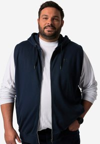 Navy zip-up hoodie with attached hood, featuring a silver zipper and a small logo. Worn over a white long-sleeve shirt.