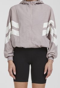 Kvinna som bär en mauve-färgad kort jacka med vita ränder på ärmarna och svarta åtsittande cykelbyxor, stående med avslappnade armar.