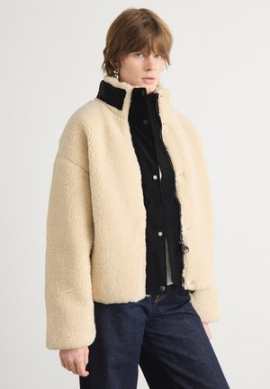 Beige faux fur jack met textuur en een zwart corduroy paneel, met een staande kraag, drukknoopsluiting en een relaxte pasvorm. Blauwe jeans maken de look compleet.