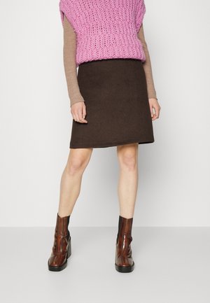 Personne portant un pull sans manches en tricot épais rose, un haut beige à manches longues, une jupe marron et des bottines marron à talons, debout devant un fond blanc.