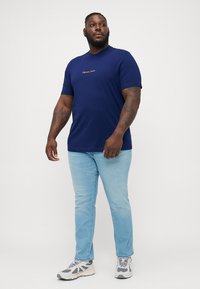 Camiseta de manga corta azul marino con un logo naranja, combinada con jeans de denim azul claro y zapatillas grises.