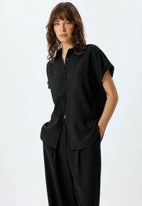 Camisa negra de manga corta y botón en tejido texturizado, con un corte relajado y mangas enrolladas, combinada con pantalones negros de cintura alta.