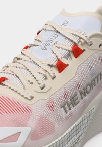 Gros plan sur une chaussure The North Face beige et blanche avec des passants de lacets rouges et des lacets blancs texturés sur une tige en tissu mesh.