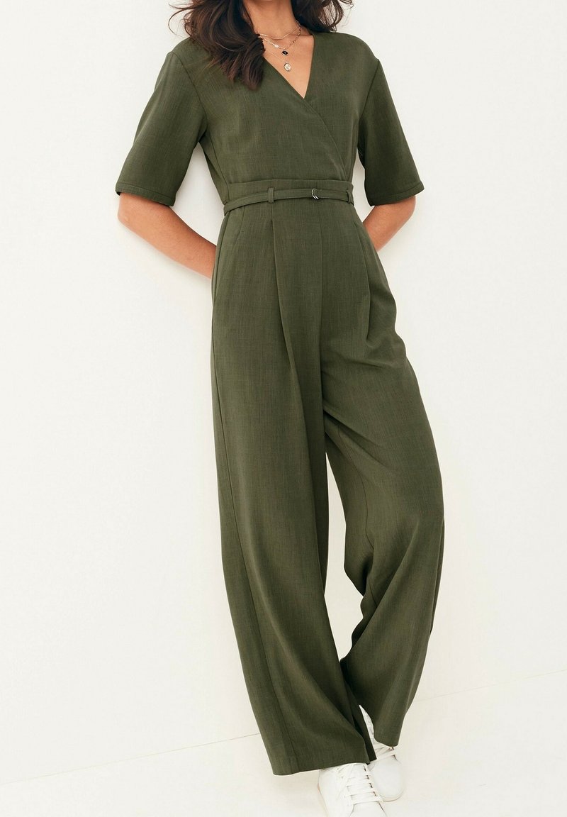 Femme portant une combinaison olive ceinturée, avec jambes larges et manches trois-quarts, associée à des baskets blanches, debout contre un mur blanc.