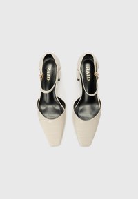 Beige nilon slingback dengan tekstur buaya, ujung runcing, dan detail logam kecil; insole hitam dengan merek "RAID".