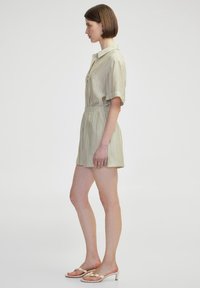 Robe chemise courte à manches courtes, verte claire, en tissu texturé, avec un col et une taille élastique. Associée à des sandales à talons beiges.
