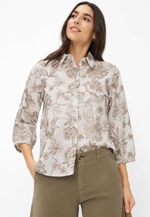 Frau trägt ein beiges Hemd mit braunen Blumenmustern und hochgekrempelten Ärmeln, kombiniert mit olivgrünen, hoch taillierten Hosen.