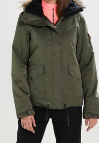 Veste isolante vert olive avec capuche noire, bordure en fourrure, fermeture éclair à l'avant et deux poches avant. Dispose de cordons ajustables à l'ourlet.