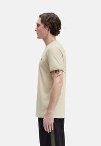 Camiseta de algodón beige con cuello redondo, mangas cortas y ribete marrón en el cuello y las mangas, que presenta un pequeño logo en el pecho.
