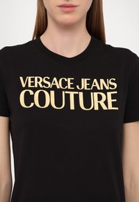 Juodas medvilnės marškinėliai su dideliu auksiniu užrašu "VERSACE JEANS COUTURE" per krūtinę. Apvalia kaklo iškirpte ir trumpomis rankovėmis.
