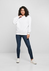 Balta sportinė palaidinė su briaunuotomis rankogaliais ir apačia, derinama su tamsiai mėlynais siaurais džinsais ir baltais sportbačiais. Modelis turi garbanotus plaukus ir stovi tvirtai.