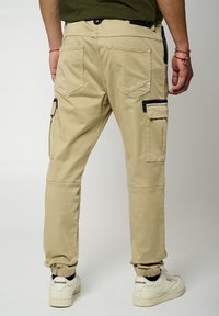 Pantalons cargo beiges avec poches latérales, poignets élastiques et texture lisse. Comprend deux poches arrière, des coutures contrastées et une coupe décontractée.