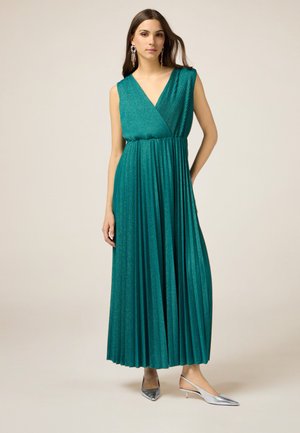 Teal pleated maxi dress with a deep V-neckline and sleeveless design, made from shimmering fabric. Silver pointed-toe heels complement the outfit.