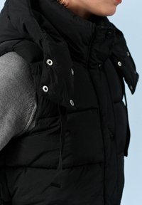 Sort puffervest med høj krave, der har store knapper, trykknaplukninger og justerbare stropper på skuldrene. Struktureret stof.