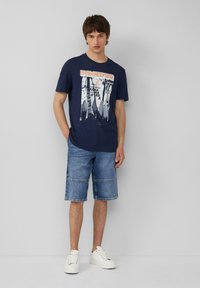 Marineblauwe katoenen t-shirt met een grafische print en tekst, gecombineerd met denim shorts en witte sneakers. Eenvoudige, casual outfit.