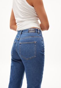 Blå denimjeans med hög midja, med bakfickor och en mörk etikett vid midjebandet. Slät textur och klassisk passform.